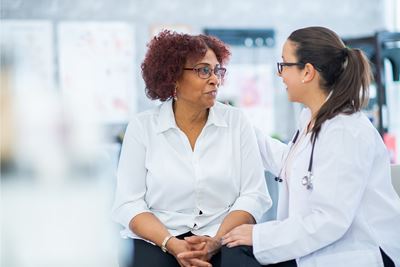 A patient -- a woman with a glasses, auburn hair and white top -- converses with a medical professional -- a woman with glasses, a clinical coat and a stethoscope around her neck. Both appear to be seated. The professional woman has a hand on the patient's shoulder."