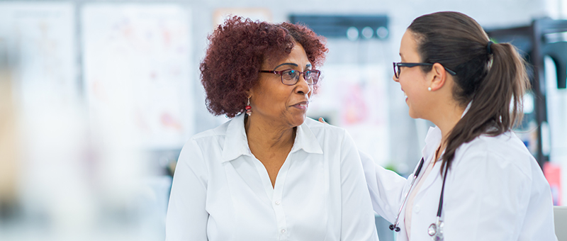A patient -- a woman with a glasses, auburn hair and white top -- converses with a medical professional -- a woman with glasses, a clinical coat and a stethoscope around her neck. Both appear to be seated. The professional woman has a hand on the patient's shoulder.